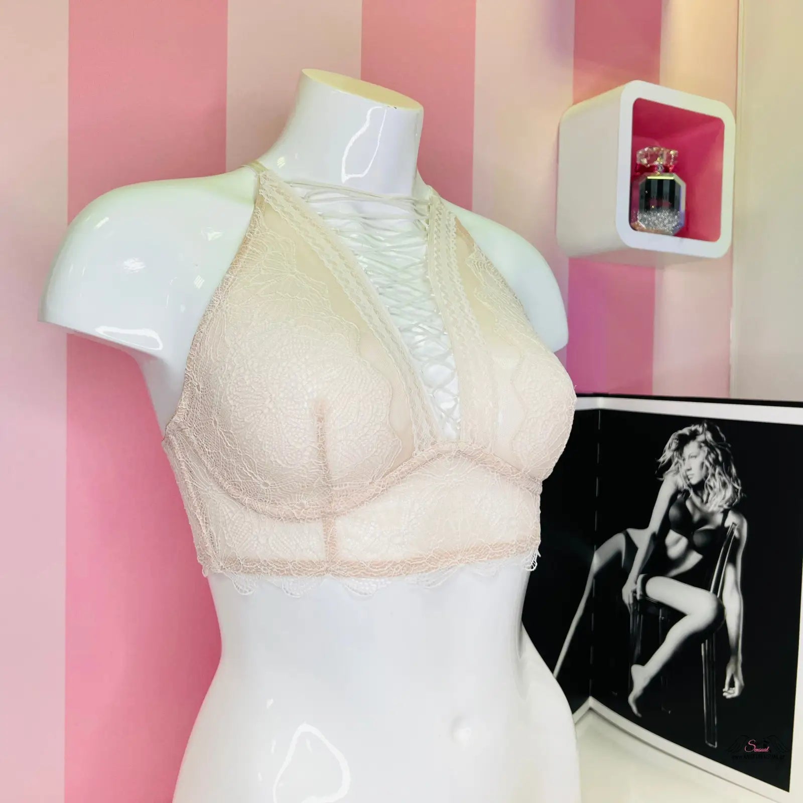 Arafovaná figurína s bílou podprsenkou Victoria’s Secret a překříženými ramínky na růžové pruhované zdi
